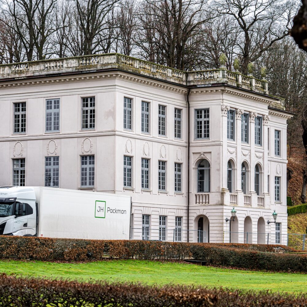 En hvid klassicistisk bygning med detaljerede gesimser og store vinduer danner baggrund for en JH Packman-lastbil, der holder parkeret ved ejendommen. Bygningens facade bærer præg af patina, mens de omkringliggende træer uden blade indikerer en vinterperiode.