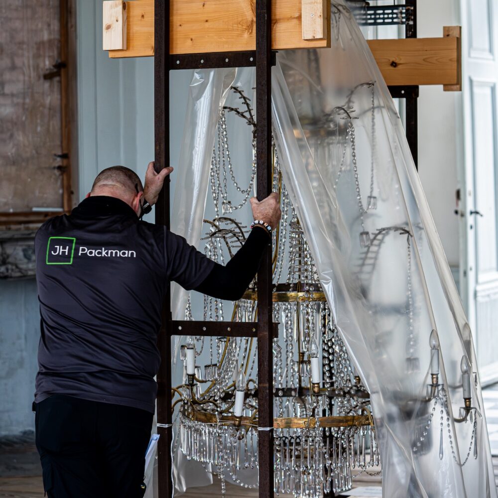 En person i en mørk jakke med “JH Packman”-logo ses bagfra, mens han forsigtigt skubber en stor lysekrone, der er monteret i en metal- og trækonstruktion. Lysekronen er delvist dækket af plast for at beskytte de mange hængende krystalprismer. Rummet har høje paneldøre, et gammelt marmorkamin og et ternet gulv, hvilket antyder en historisk bygning.