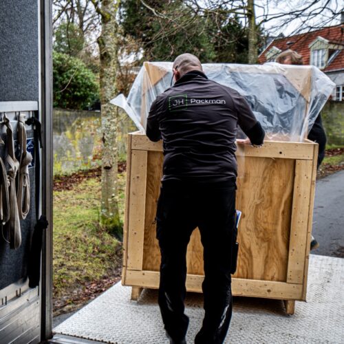 En trækasse med en lysekrone skubbes op ad en rampe ind i en transportvogn. Indvendigt i vognen ses fastspændingsstropper, der sikrer en stabil transport. I baggrunden ses en have og et hus med rødt tegltag, hvilket antyder en forsendelse fra en herskabelig ejendom.