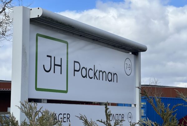 Skilt med JH Packman-logo ved virksomhedens adresse, med blå himmel og bygninger i baggrunden.